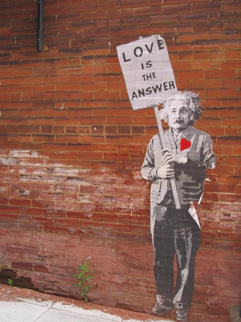 Street-art paste-up on a brick wall: Albert Einstein with a red heart on his chest holds a sign that reads Love is the answer